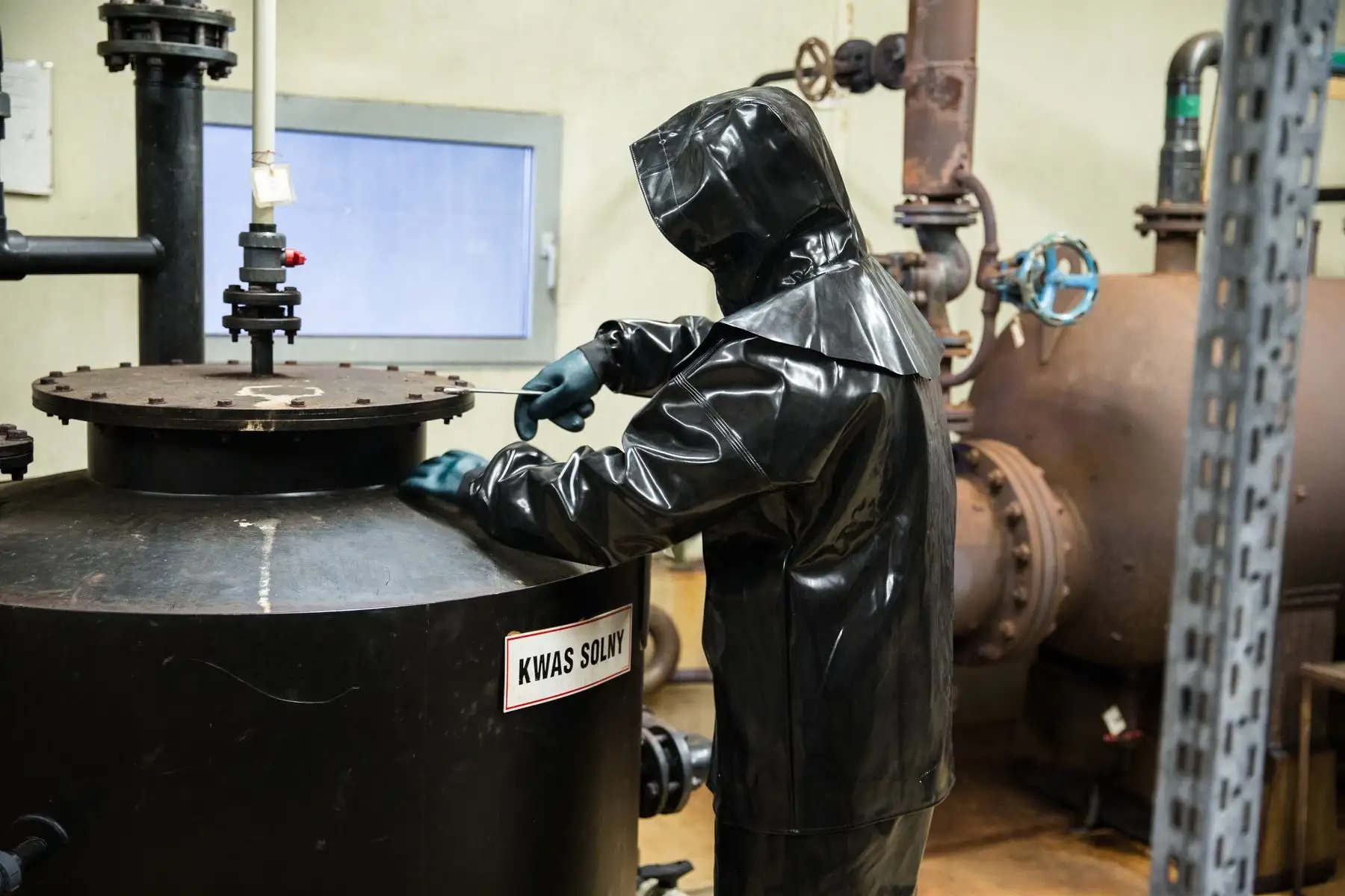 Chemikant in Chemikalienschutzanzug mit Kapuze und Handschuhen arbeitet an einem Behälter für Salzsäure in einer Produktionsanlage.