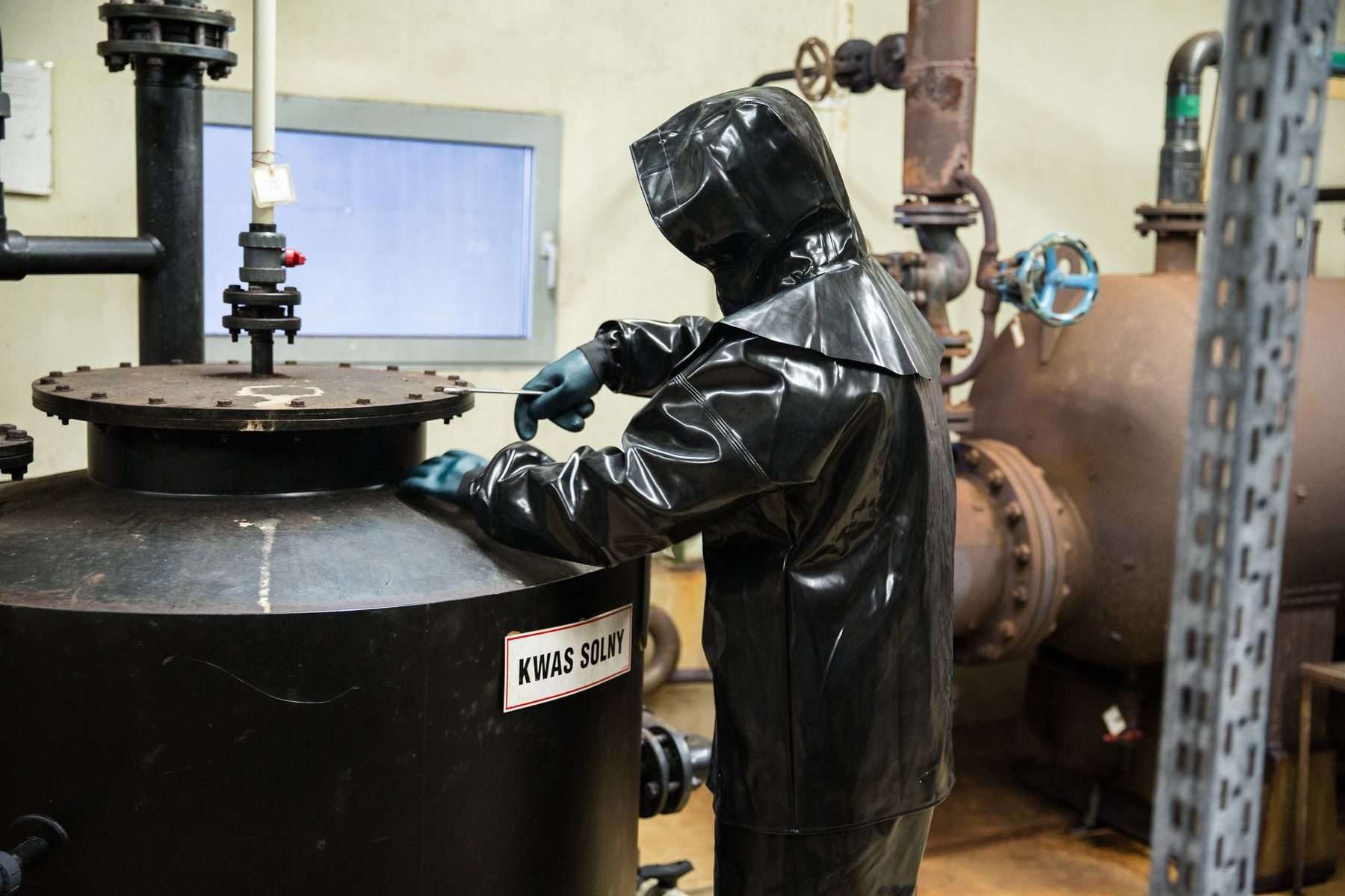 Chemikant in Chemikalienschutzanzug mit Kapuze und Handschuhen arbeitet an einem Behälter für Salzsäure in einer Produktionsanlage.