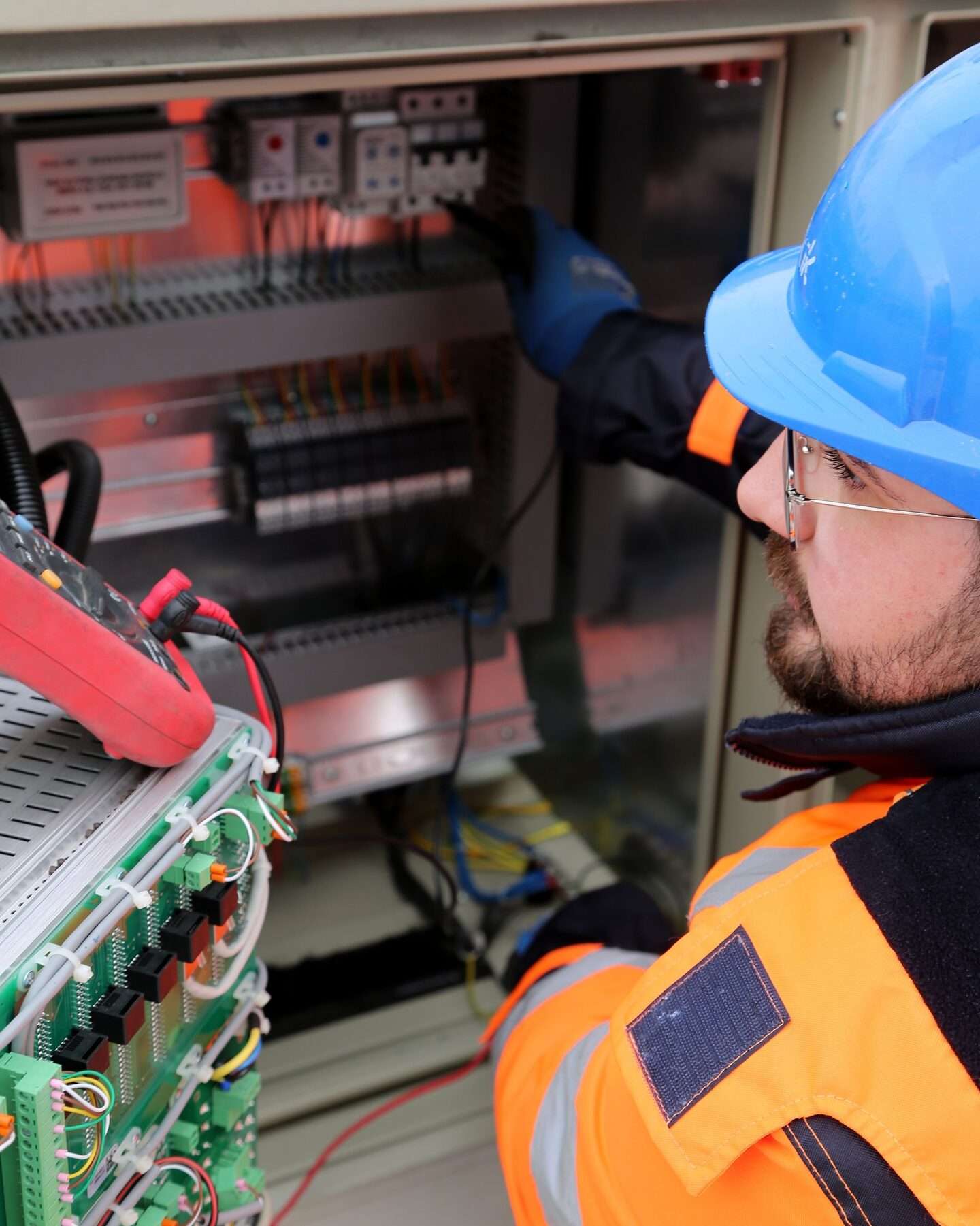 Elektriker mit Schutzhelm, Schutzbrille und Warnjacke arbeitet mit Strommesszange in einem Schaltschrank.