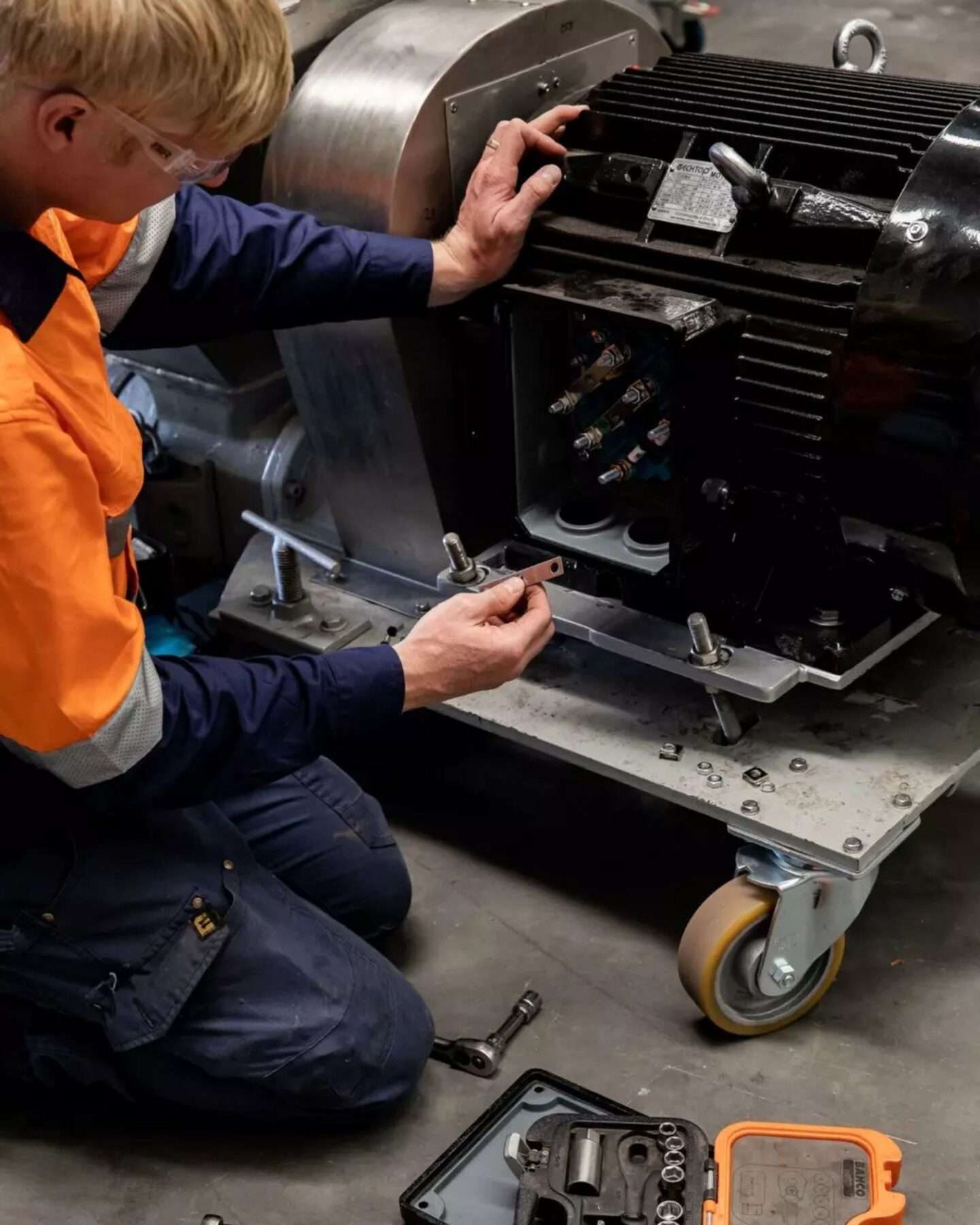 Mechaniker mit Schutzbrille kniet an einem Elektromotor auf einem Montagewagen und legt ein Ausrichtblech unter den Motorfuß, daneben ein offener Steckschlüsselkasten.