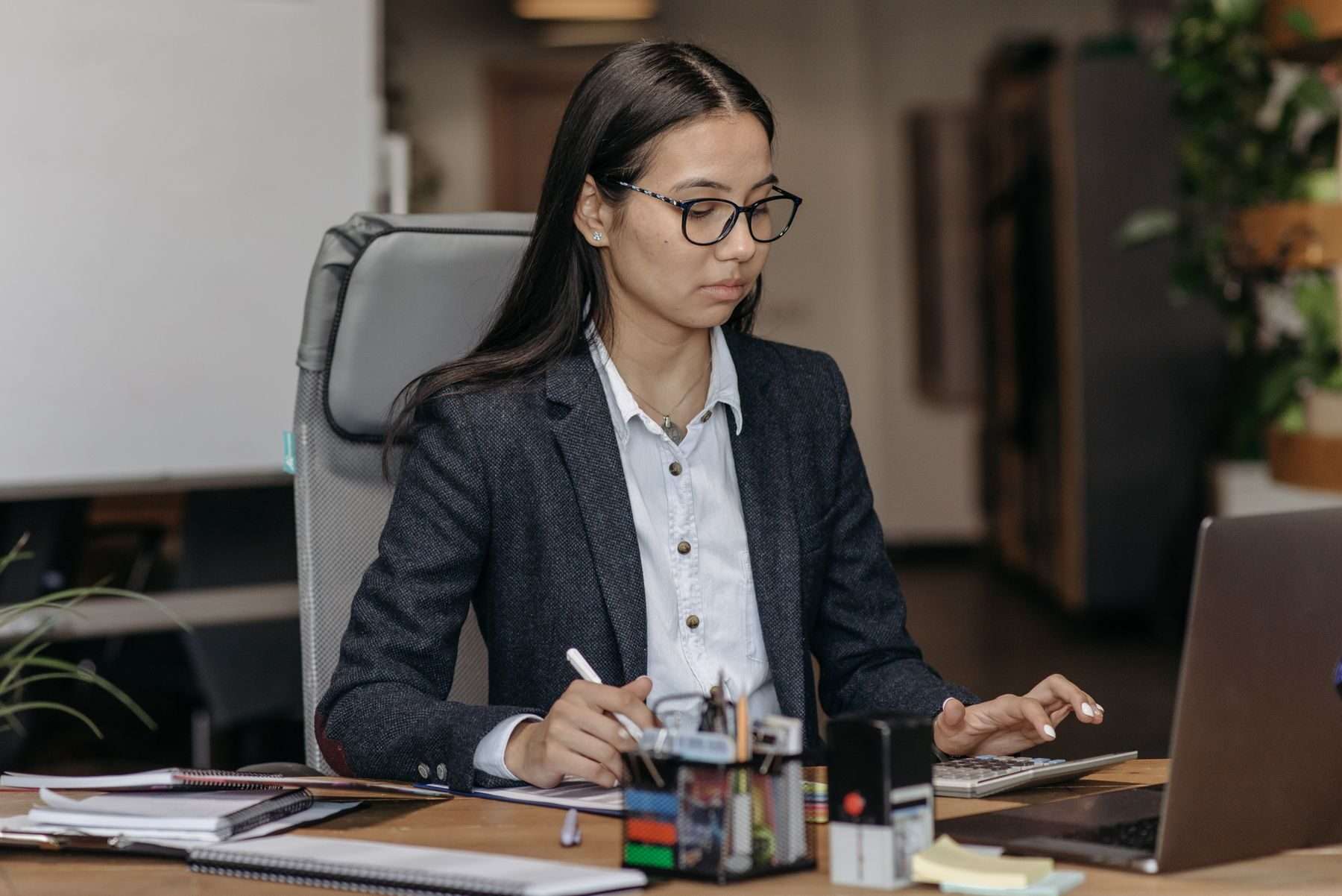 Junge Frau mit Brille im Büro am Schreibtisch, rechnet mit dem Taschenrechner und notiert, daneben Stempel und Laptop.