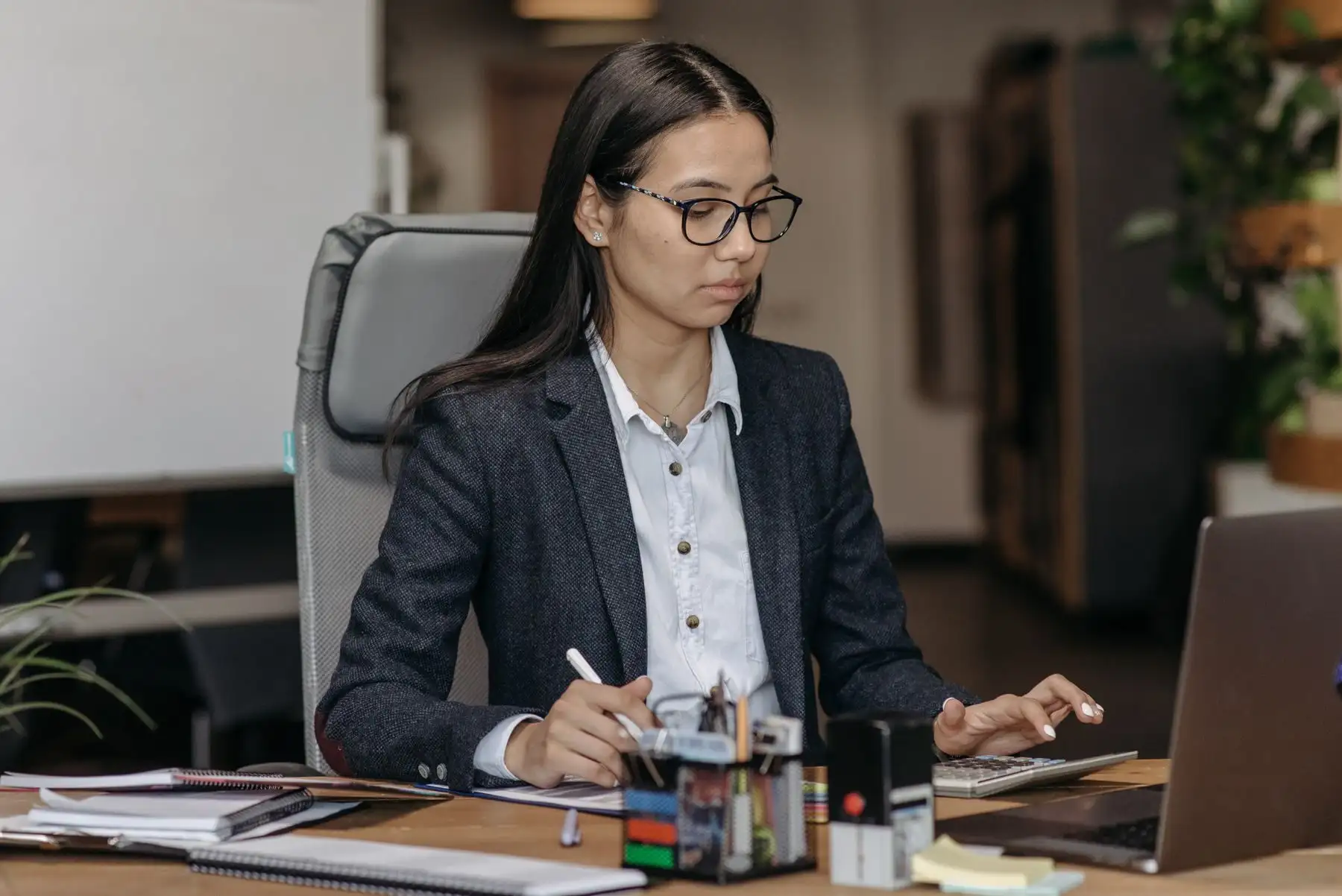 Junge Frau mit Brille im Büro am Schreibtisch, rechnet mit dem Taschenrechner und notiert, daneben Stempel und Laptop.