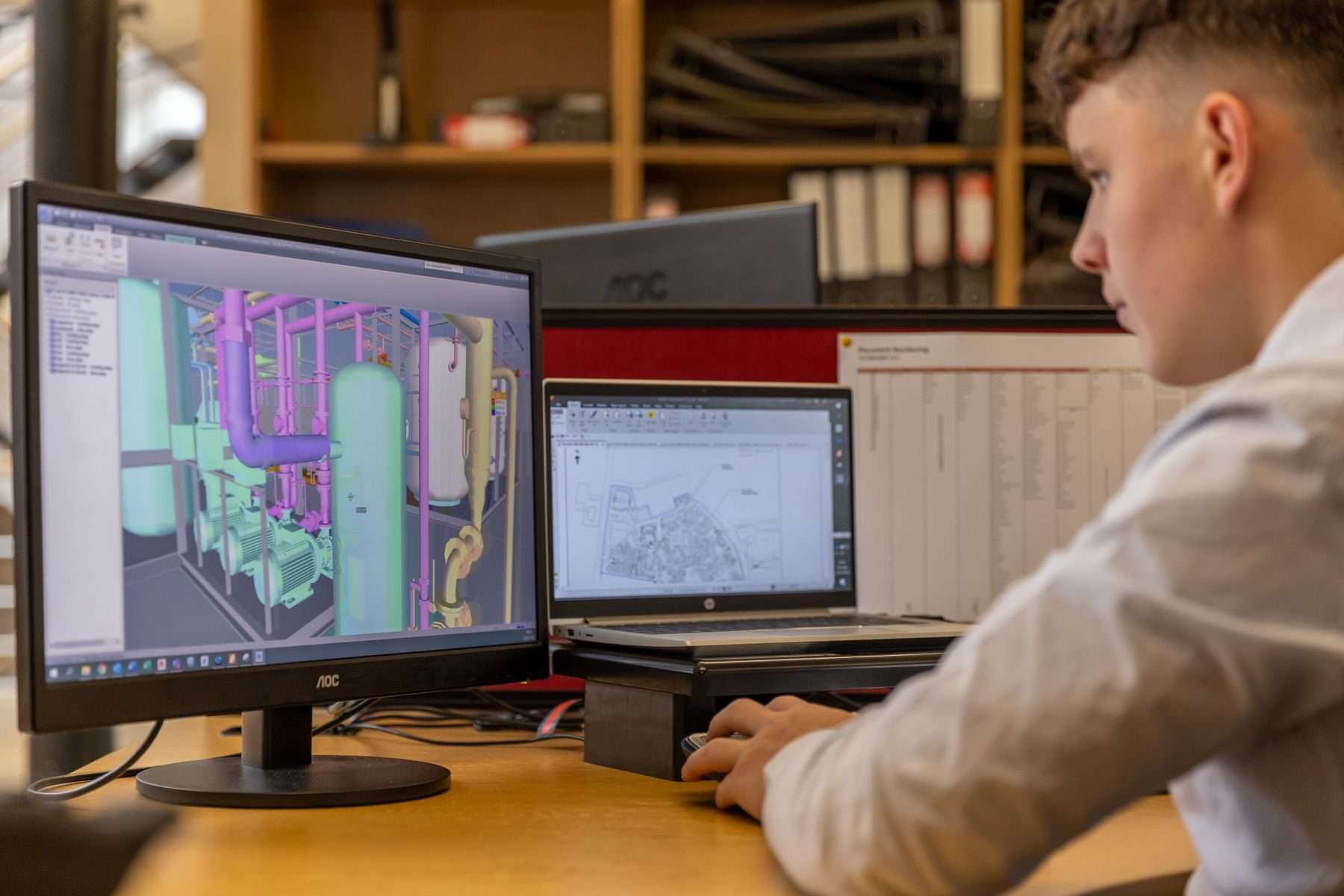 Junger Mann im Büro plant am Bildschirm eine Anlage mit Rohrleitungen und Behältern im 3D-Modell, daneben ein Lageplan auf dem Laptop.