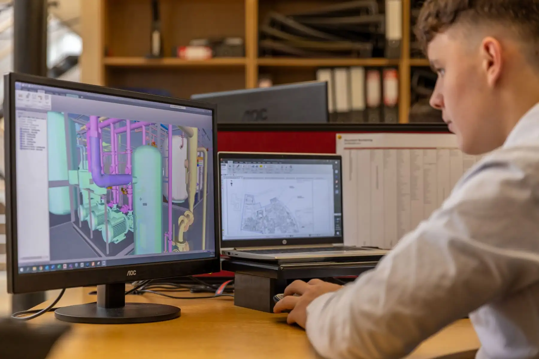 Junger Mann im Büro plant am Bildschirm eine Anlage mit Rohrleitungen und Behältern im 3D-Modell, daneben ein Lageplan auf dem Laptop.