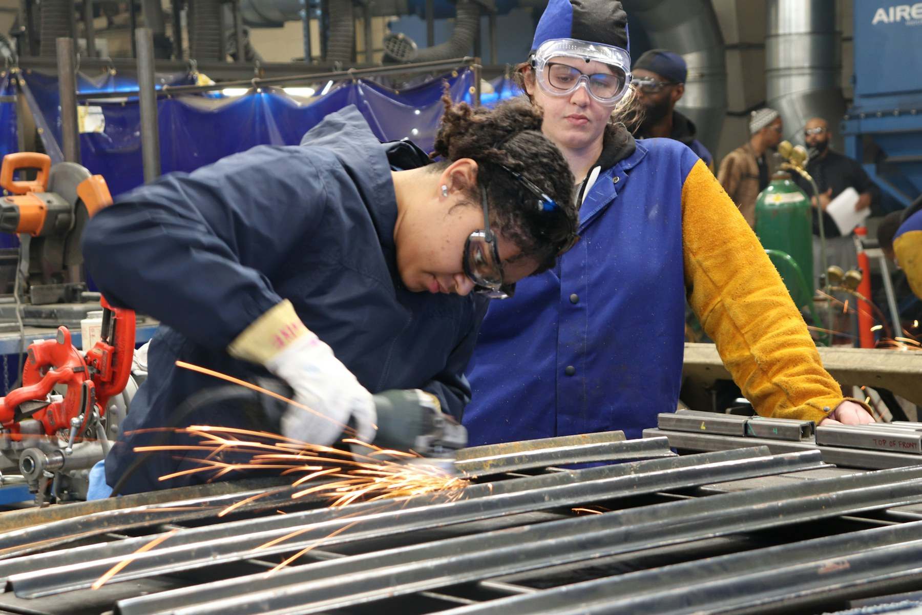 Auszubildende mit Schutzbrille schleift in der Werkstatt ein Metallteil, eine Kollegin schaut zu